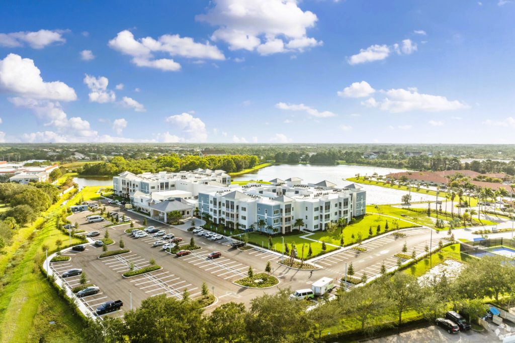 Bird's-eye view of Aviva Port St. Lucie, a luxury independent living community for adults 55+ by ST Real Estate, showcasing modern low-rise buildings surrounded by well-maintained greenery, a scenic lake, and ample parking under a bright, sunny sky.